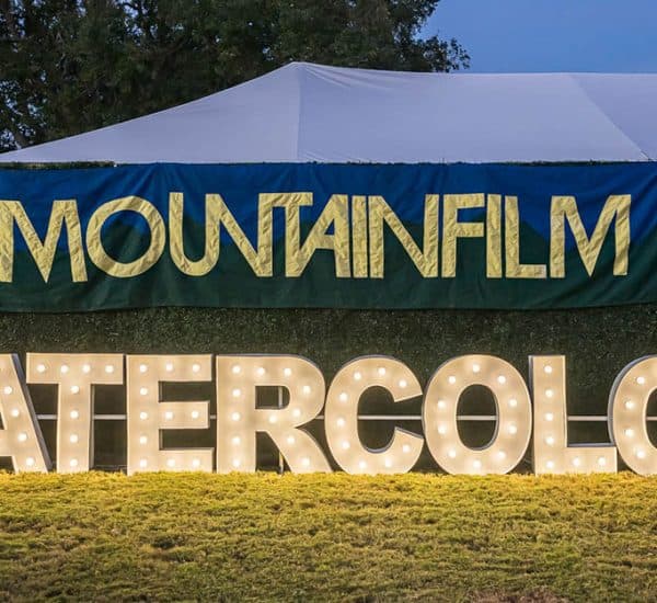 A large lit sign reading "WATERCOLOR" stands on grass in front of a banner that says "MOUNTAINFILM" with a white tent behind it.