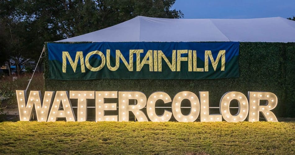 A large lit sign reading "WATERCOLOR" stands on grass in front of a banner that says "MOUNTAINFILM" with a white tent behind it.