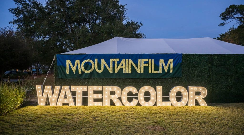 A large illuminated "WATERCOLOR" sign stands on grass in front of a tent with a "MOUNTAINFILM" banner during an outdoor event at dusk.