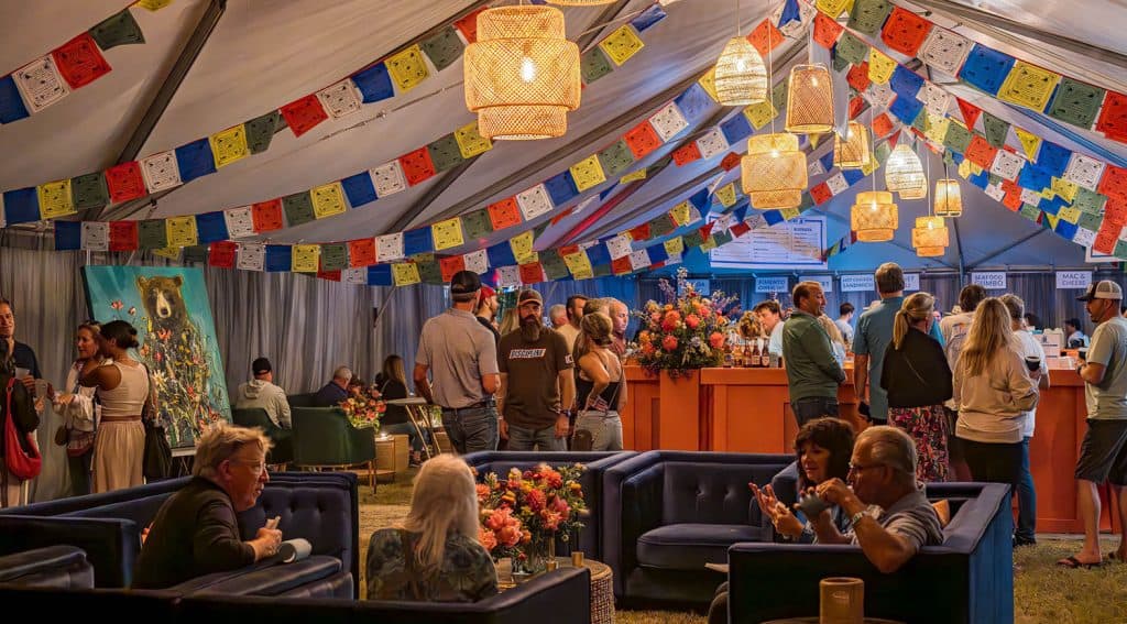 People gather inside a decorated tent with colorful banners, lounge seating, and hanging lights, some sitting and others standing at a bar or counter.