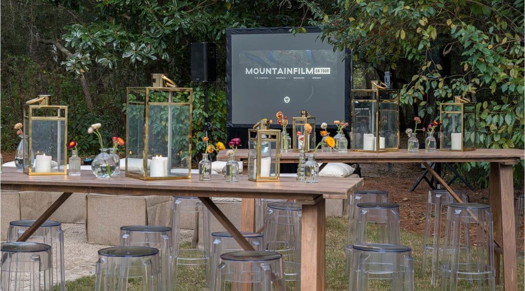 Outdoor event setup with wooden tables, glass lanterns, small flower vases, clear chairs, and a screen displaying "Mountainfilm on Tour" surrounded by greenery.