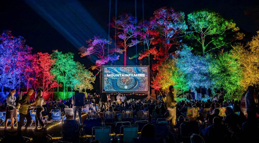 Outdoor nighttime film screening with a crowd seated in front of a large screen; surrounding trees are brightly lit with multicolored lights.