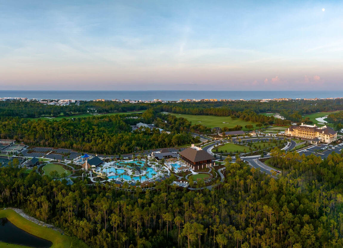 Aerial view of a large resort complex with multiple pools, buildings, and parking areas, surrounded by dense greenery under a clear sky.