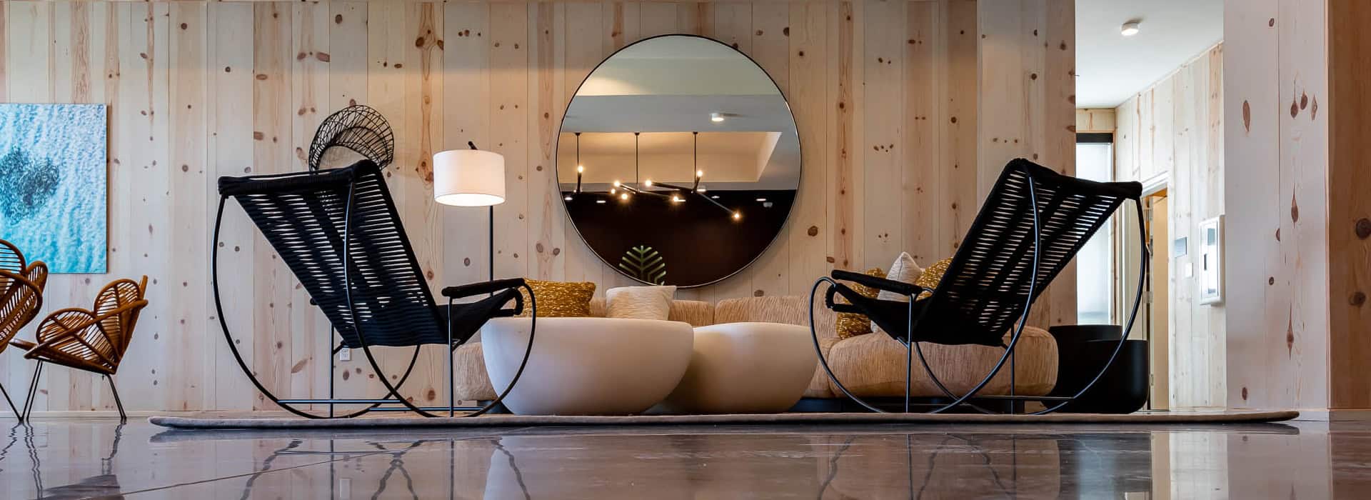 The lobby of The Lodge 30A in Greenway Station along Hwy 30A. Modern living room with two black rocking chairs, a beige sofa, round white coffee tables, a large round mirror, and light wood panel walls.