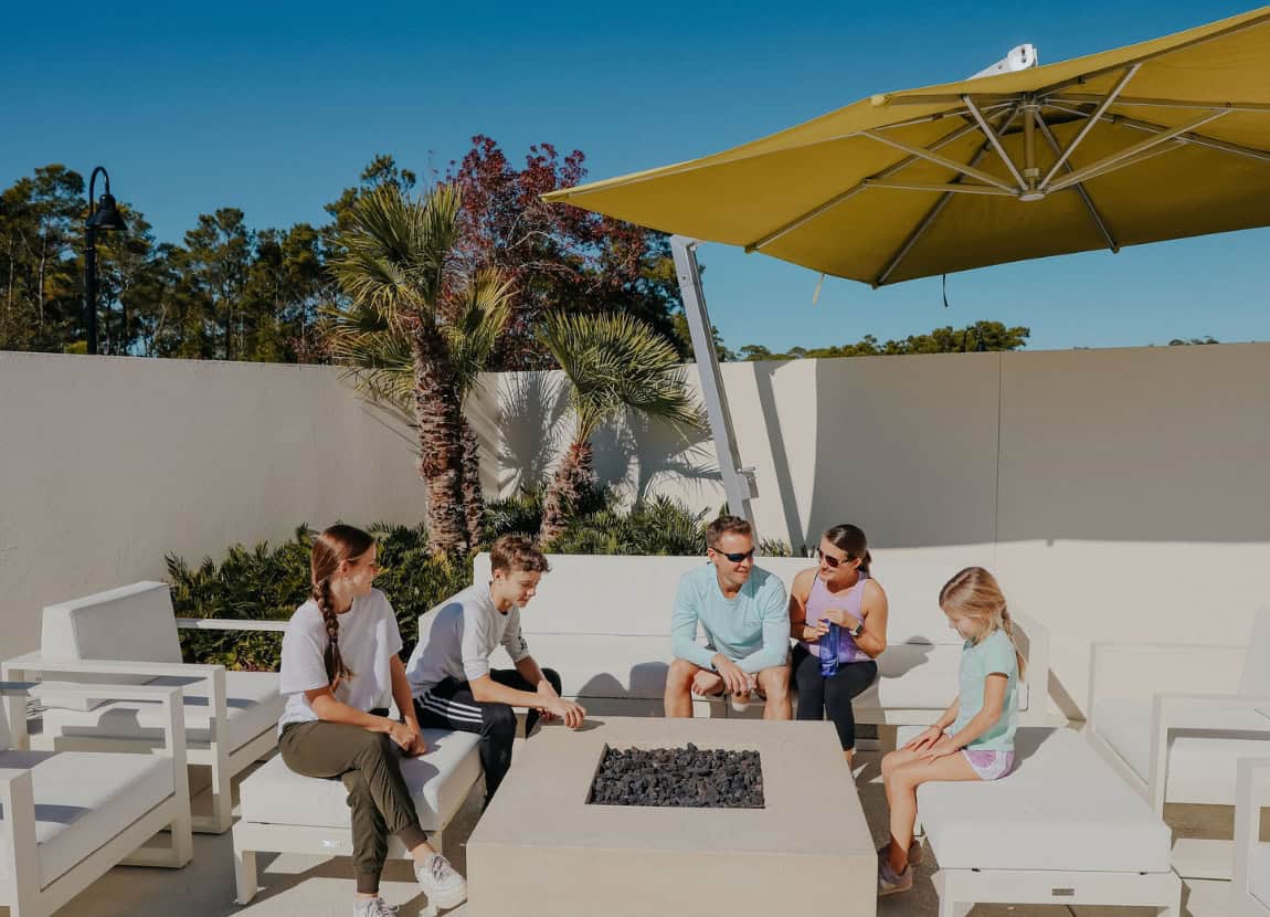 Five people sit around a modern outdoor fire pit under a large yellow umbrella, with palm trees and white walls in the background.