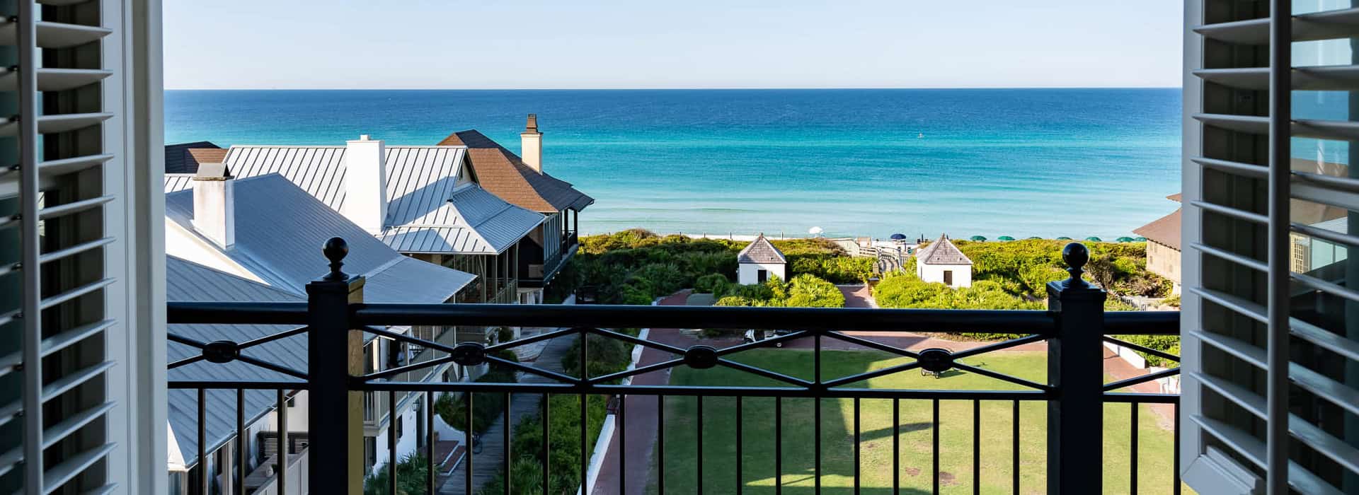 View from a balcony overlooking rooftops, green lawns, and pathways leading to a beach with clear blue water under a clear sky.