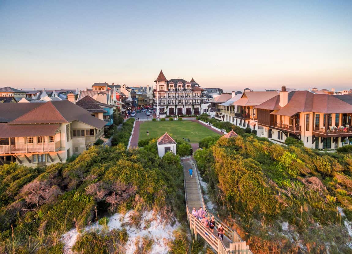 Aerial view of a coastal town with a large, ornate building in the center, surrounded by smaller houses, greenery, and a boardwalk leading to the beach.