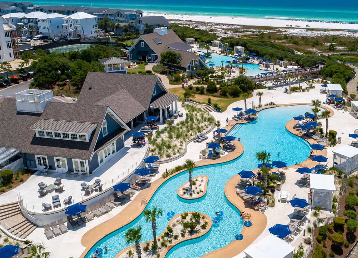 Aerial view of the Watersound Beach Club with winding outdoor pools, sun loungers, cabanas, and several buildings near a sandy beach and turquoise ocean.