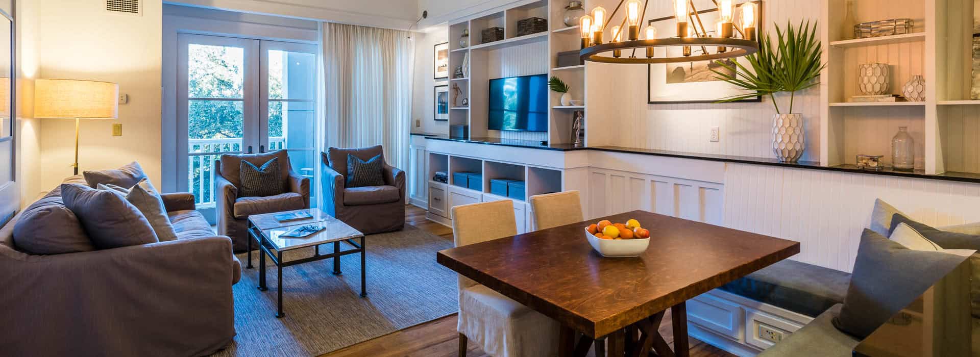 Watersound Inn Living area. A contemporary living and dining area with a wooden table, gray couches, built-in shelves, wall-mounted TV, and large window with sheer curtains.