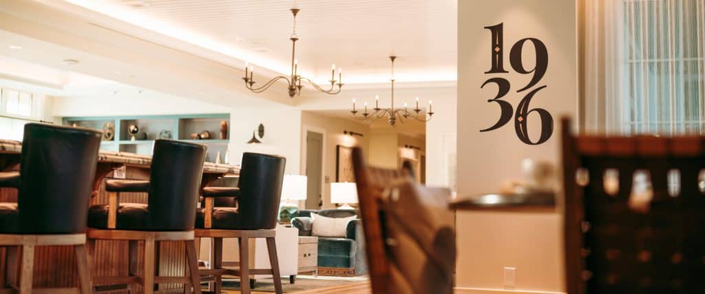 Upscale lounge area with wooden bar stools, a bar counter, modern chandeliers, and the numbers "1936" displayed on a wall.