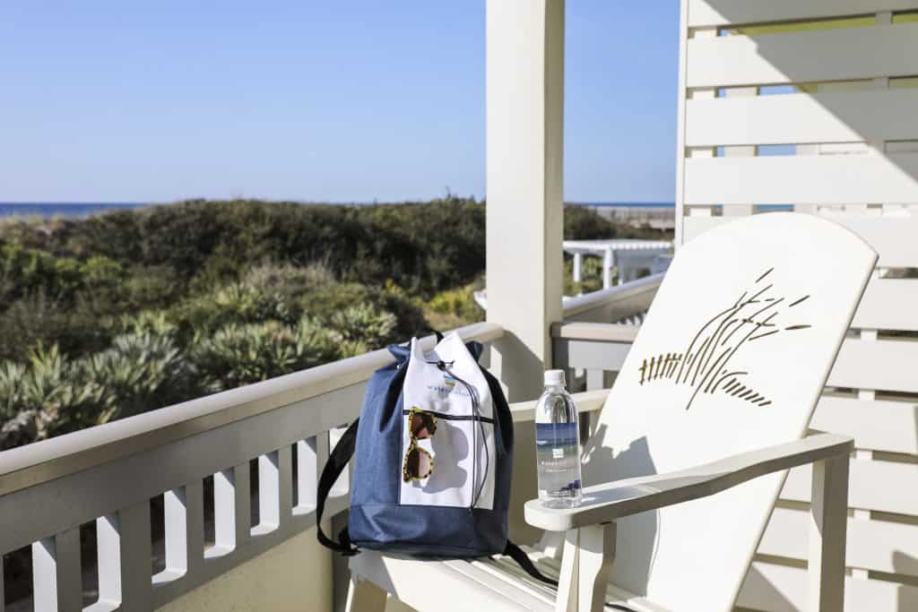 A blue and white backpack, sunglasses, and a water bottle rest on a white chair on a porch overlooking greenery and the ocean.