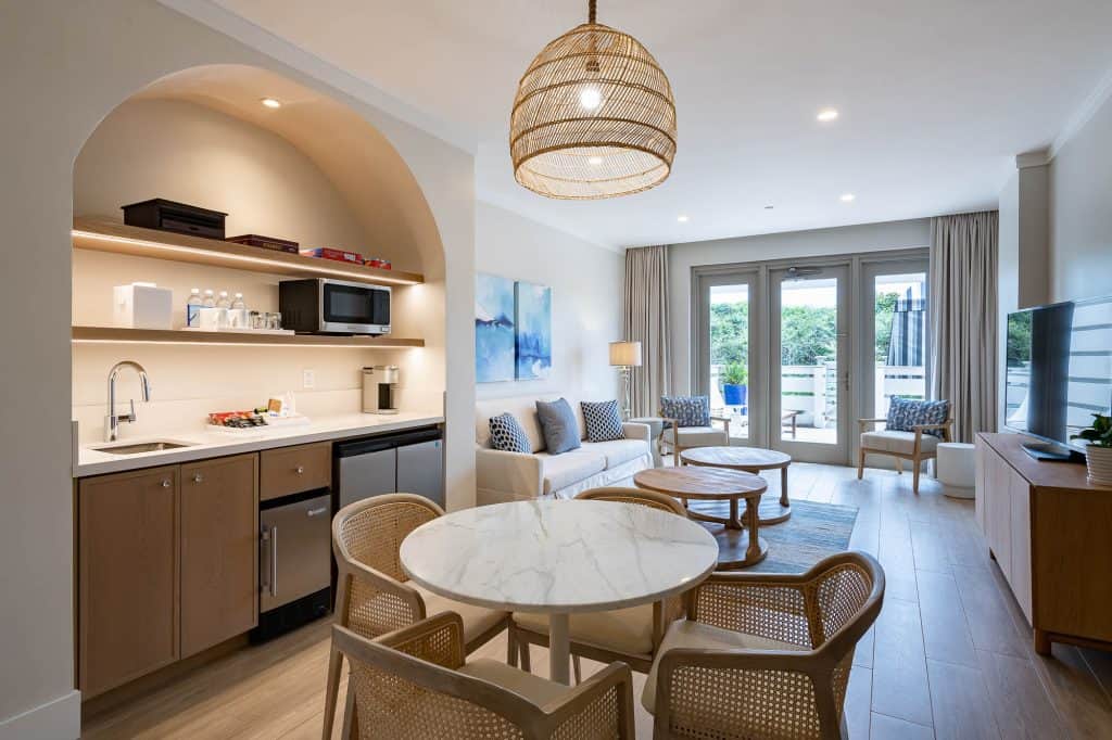 Modern open-plan hotel suite with a kitchenette, dining table, seating area, TV, and large windows leading to a balcony with views of greenery.