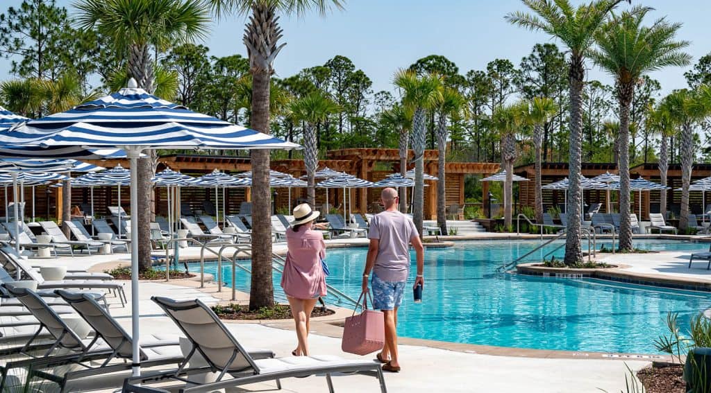 Two people walk beside a resort swimming pool lined with lounge chairs and blue-and-white umbrellas surrounded by palm trees.