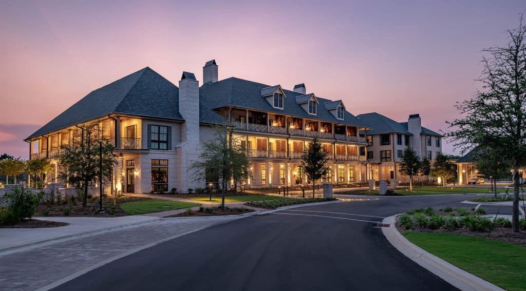 A large, well-lit building with multiple balconies and dormer windows at sunset, surrounded by landscaped grounds and a paved driveway.