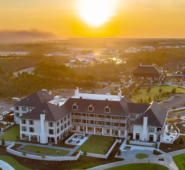 Aerial view of a large, elegant building surrounded by landscaped grounds, roads, and golf courses at sunset with a hazy sky in the background.