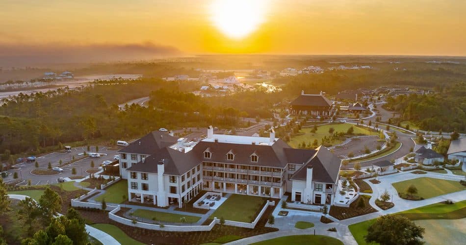 Aerial view of a large, elegant building surrounded by landscaped grounds, roads, and golf courses at sunset with a hazy sky in the background.