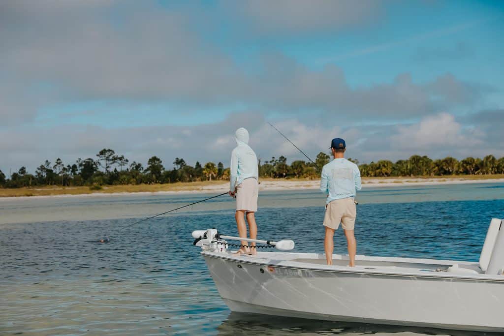 Two people in light-colored clothes are fishing from the deck of a small boat on calm water near a shoreline with trees under a partly cloudy sky.
