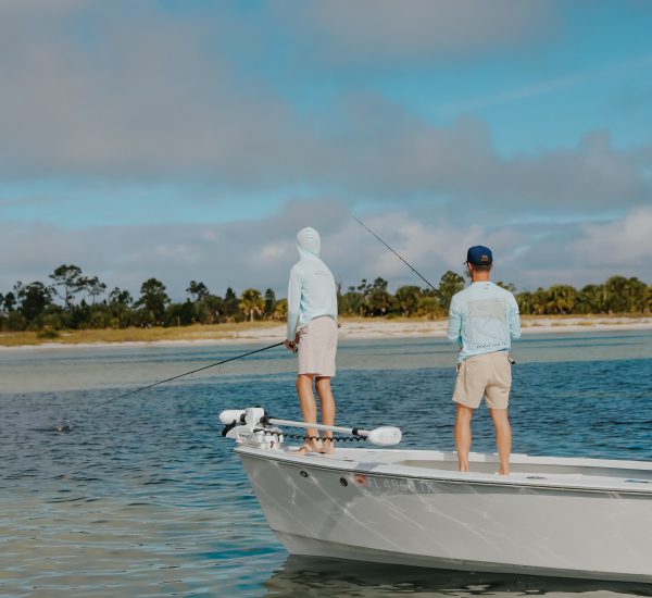 Two people in light-colored clothes are fishing from the deck of a small boat on calm water near a shoreline with trees under a partly cloudy sky.