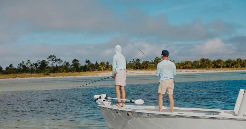 Two people in light-colored clothes are fishing from the deck of a small boat on calm water near a shoreline with trees under a partly cloudy sky.