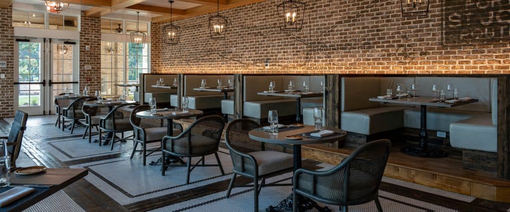 A restaurant interior with brick walls, tiled floors, booth and table seating, glassware, and natural light from large windows.