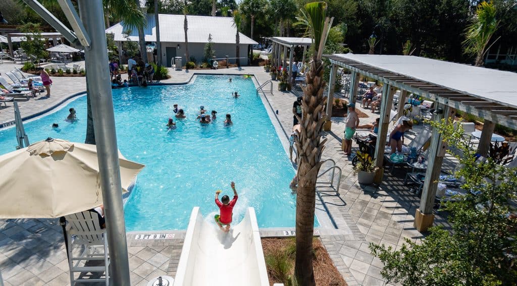 A child in a red shirt slides into a busy outdoor swimming pool, while people swim and relax on lounge chairs and under shaded areas nearby.