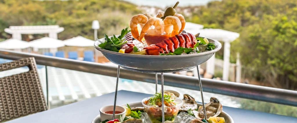 A two-tiered seafood platter with shrimp, lobster, oysters, and garnishes on ice, set on an outdoor table overlooking a pool and greenery.