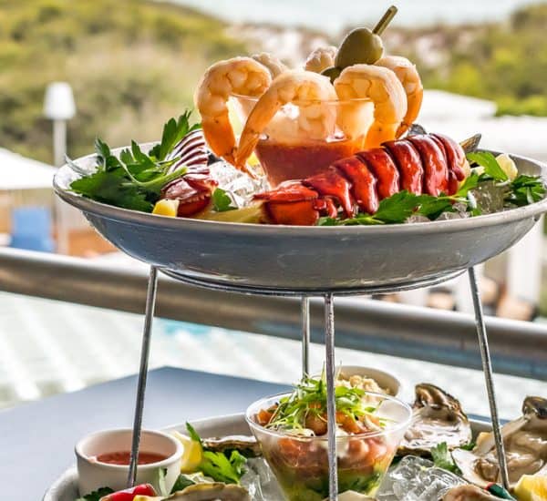 A two-tiered seafood platter with shrimp, lobster, oysters, and garnishes on ice, set on an outdoor table overlooking a pool and greenery.