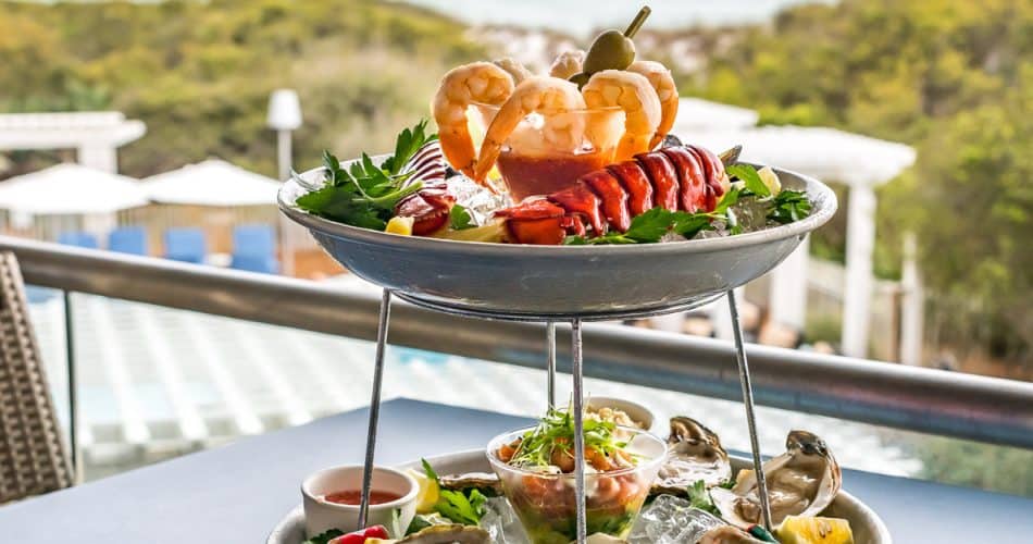 A two-tiered seafood platter with shrimp, lobster, oysters, and garnishes on ice, set on an outdoor table overlooking a pool and greenery.
