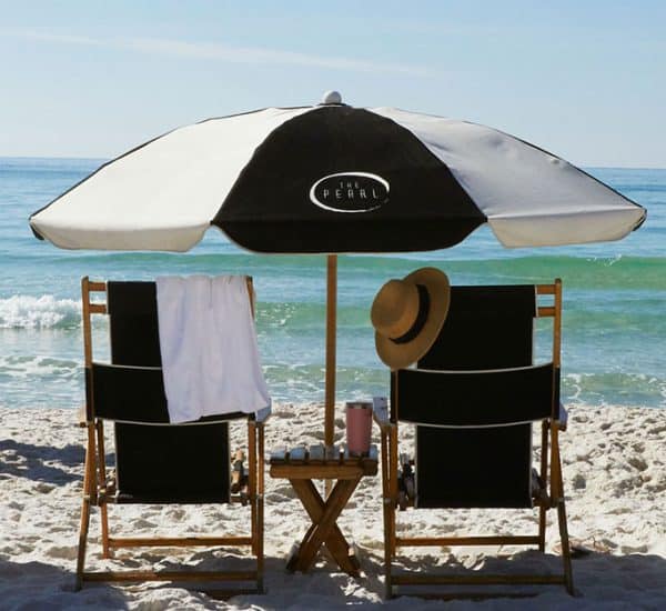 Two people walk along a sandy beach near black-and-white umbrellas and lounge chairs facing the ocean under clear blue skies.