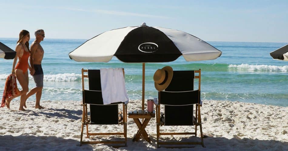 Two people walk along a sandy beach near black-and-white umbrellas and lounge chairs facing the ocean under clear blue skies.