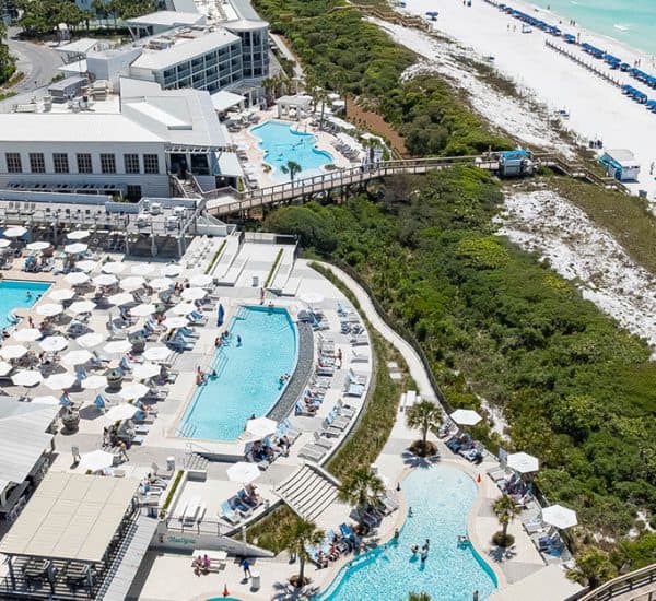 Aerial view of a beachfront resort with multiple swimming pools, sun loungers, white sand beach, turquoise water, and a boardwalk through greenery.