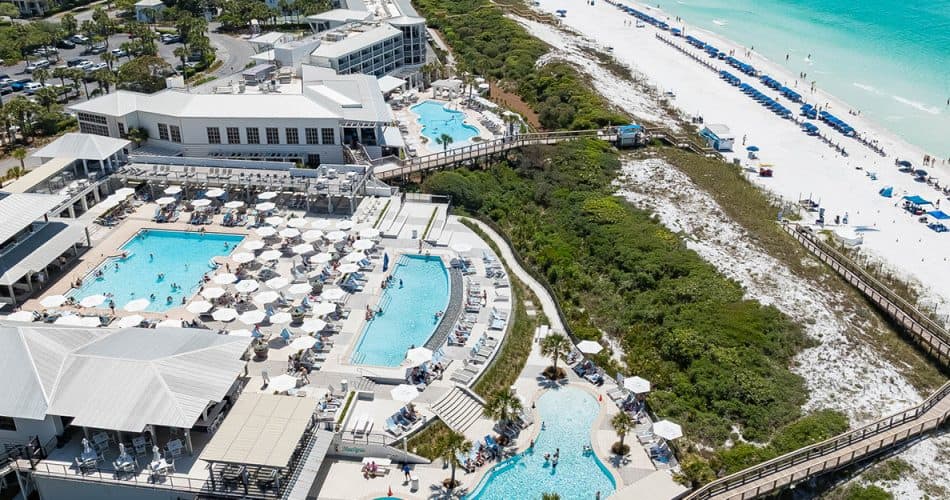 Aerial view of a beachfront resort with multiple swimming pools, sun loungers, white sand beach, turquoise water, and a boardwalk through greenery.