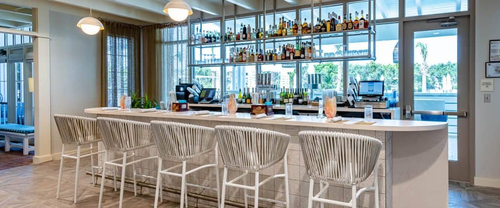 Modern bar with six white chairs, a curved counter, and shelves stocked with bottles; large windows in the background admit natural light.