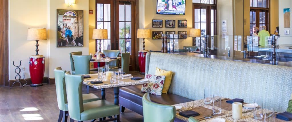A brightly lit restaurant with green chairs, cushioned booths, wall art, large windows, lamps, and a TV mounted above framed photos. Tables are set with glasses and cutlery.