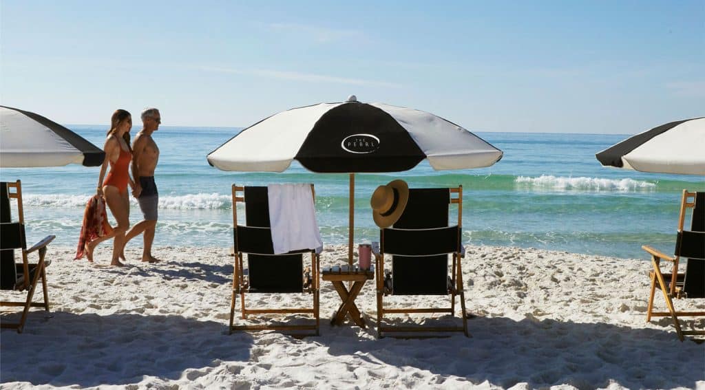 Two people walk along a sandy beach near the ocean, passing by empty lounge chairs with towels, a hat, and umbrellas.