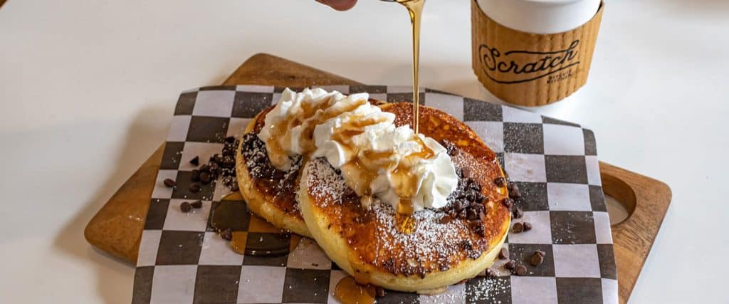 Two pancakes topped with whipped cream, chocolate chips, and powdered sugar are being drizzled with syrup, with a cup of coffee labeled "Scratch" in the background.
