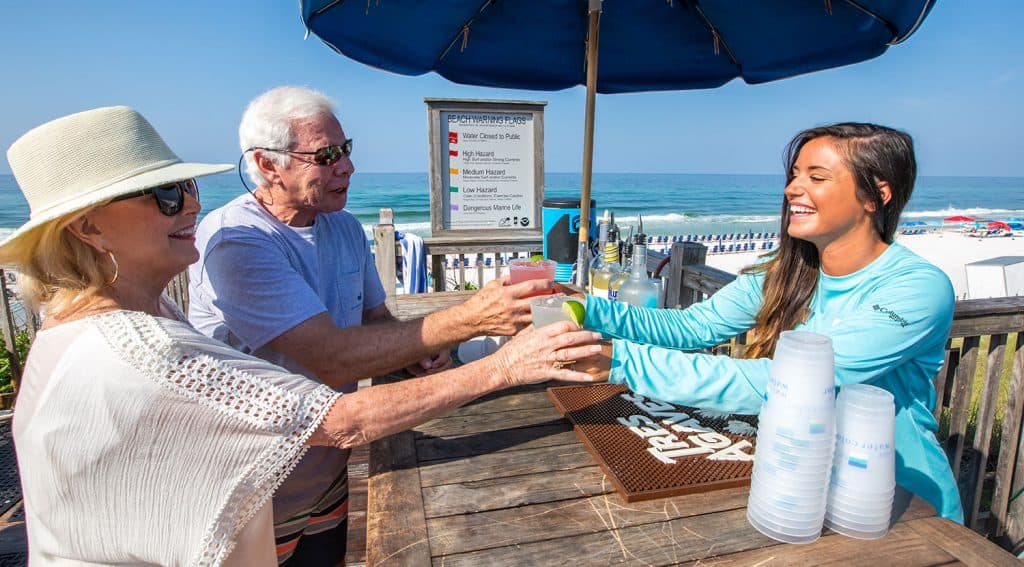 A smiling bartender hands drinks to an older man and woman at an outdoor beachside bar, with the ocean and beach in the background.
