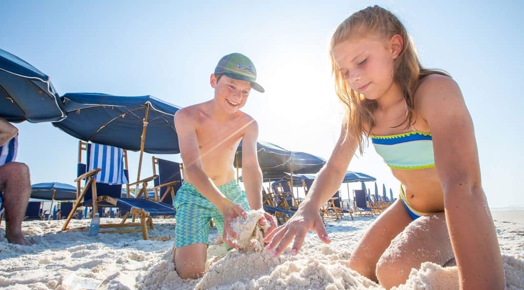 Two children in swimsuits play with sand on a sunny beach, with blue striped umbrellas and lounge chairs in the background.