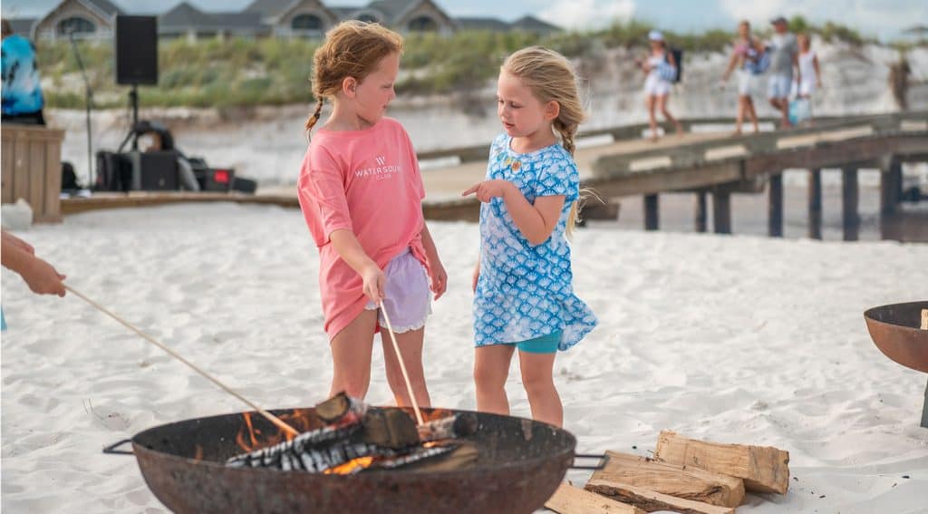 Two young girls stand on a sandy beach, roasting marshmallows over a fire pit, with more people and houses visible in the background.