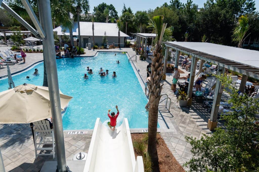 A child in a red shirt slides down a white water slide into a pool, while people swim and relax on lounge chairs around the pool area.