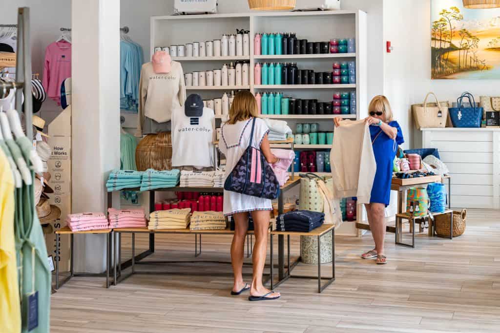 Two women shop in a boutique; one examines a t-shirt while the other looks at a folded item. Shelves behind them display water bottles, bags, and folded clothing.
