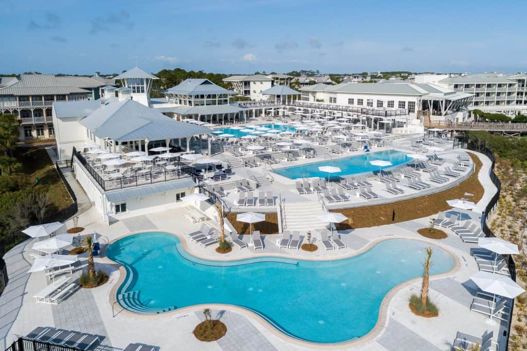 Large resort complex with multiple outdoor swimming pools, numerous white lounge chairs, and umbrellas, surrounded by modern white buildings under a clear blue sky.