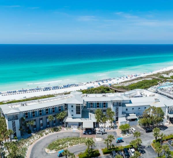 A large beachfront resort with a parking lot and palm trees sits beside a white sand beach and turquoise ocean under a clear blue sky.