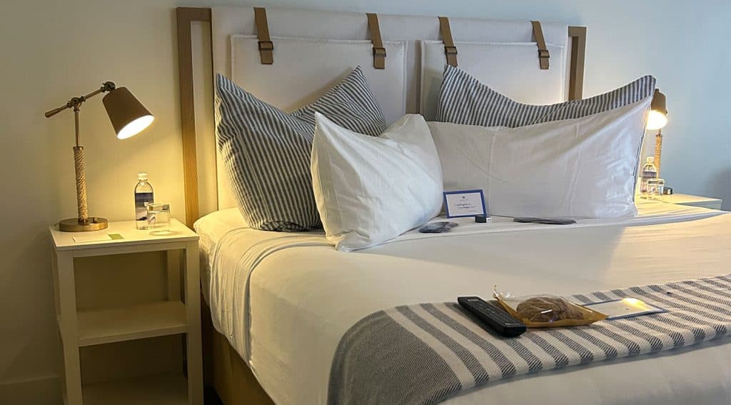 A neatly made bed with striped pillows and blanket, flanked by two nightstands with lamps, water bottles, and a TV remote with cookies placed on the bed.