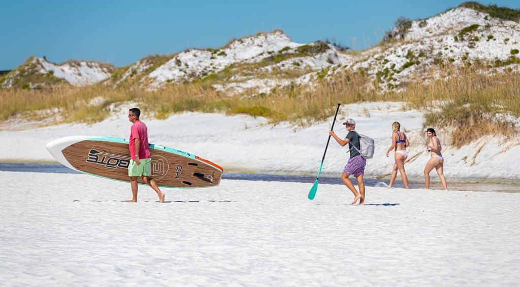Two men carry paddleboards on a sandy beach while three women walk nearby; dunes and grasses are visible in the background under a clear blue sky.