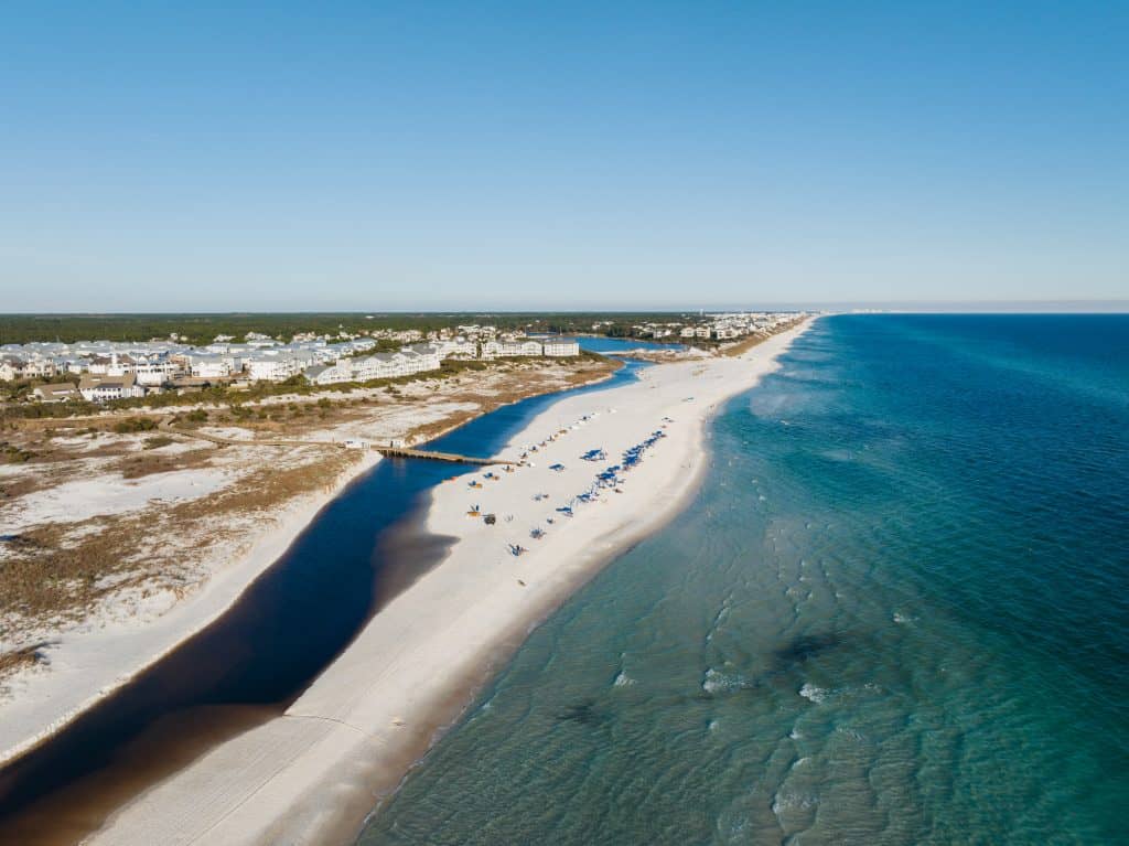 Aerial view of a sandy beach with blue umbrellas, a coastal town, a narrow inlet, and calm ocean water under a clear sky.