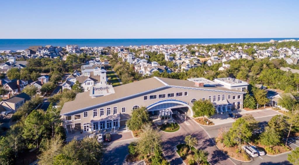 A large building with an arched entrance sits among houses and trees near the ocean under a clear blue sky.