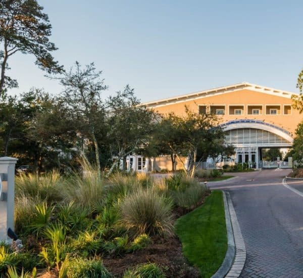 A large building with an arched entrance is seen behind landscaped greenery and a sign reading "WaterSound Beach" near a brick driveway.