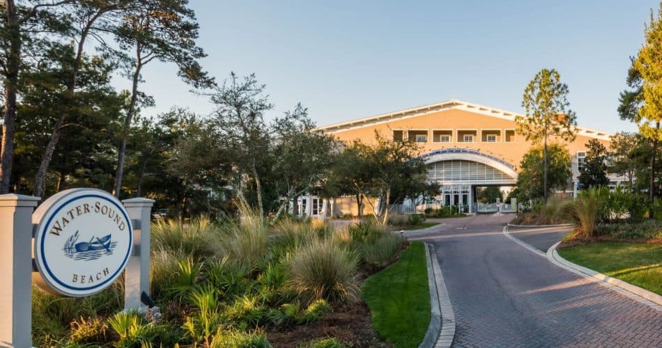 A large building with an arched entrance is seen behind landscaped greenery and a sign reading "WaterSound Beach" near a brick driveway.