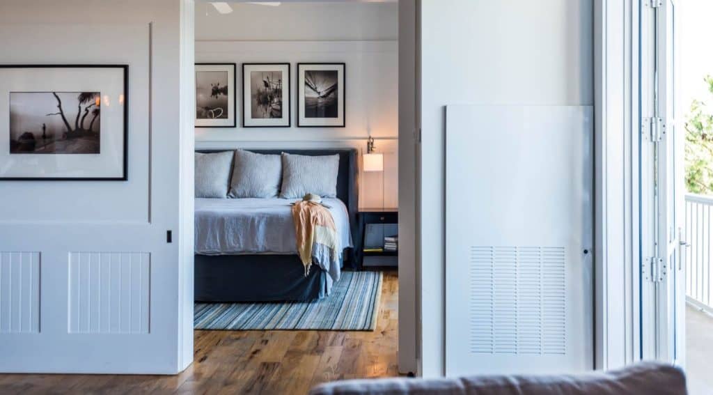 A bedroom with a neatly made bed, gray bedding, framed black-and-white photos on the wall, a nightstand with a lamp, and natural light coming from an open door.